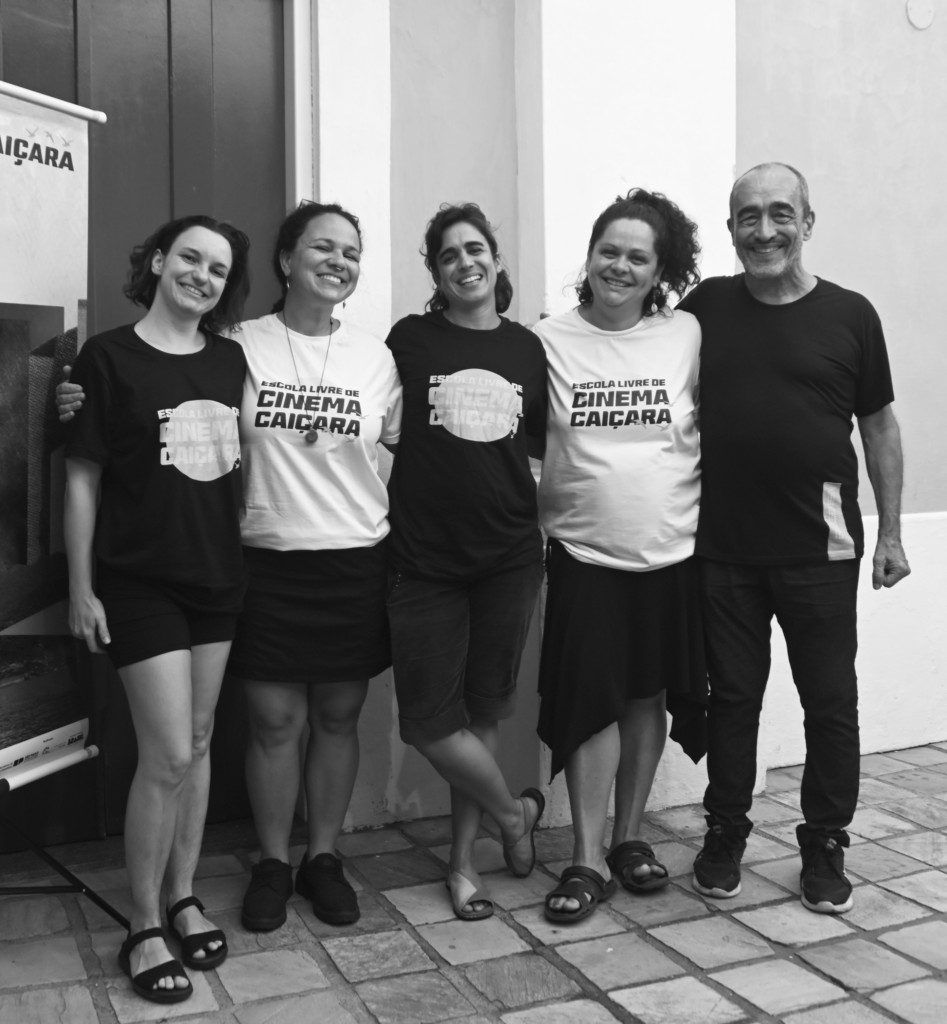 Foto em preto e branco com cinco pessoas em pé usando camisetas com o logotipo da Escola Livre de Cinema Caiçara. Eles sorriem em frente a uma parede branca. São cinco pessoas da equipe da direita para a esquerda: Mayara Mantovani, Débora Bergamini, Juliana Borges, Dyulie Ben Bolon e Francisco Cesar Filho.