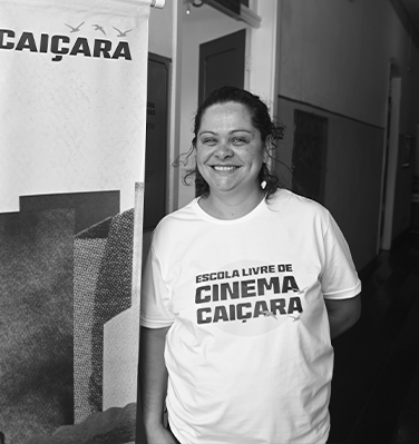 Sou uma mulher cis, parda, de 40 anos. Tenho olhos verdes e cabelos cacheados amarrados. Estou sorrindo e vestindo uma camiseta escrito Escola Livre de Cinema Caiçara.