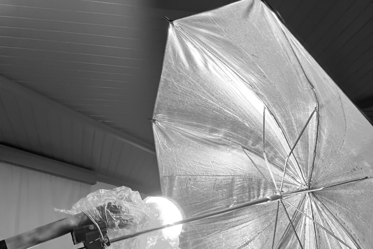 uma fotografia preto e branco de um guarda-chuva prateado, que parece ser um equipamento de iluminação usado em fotografia ou filmagem. Ele está aberto e inclinado, com as hastes visíveis.