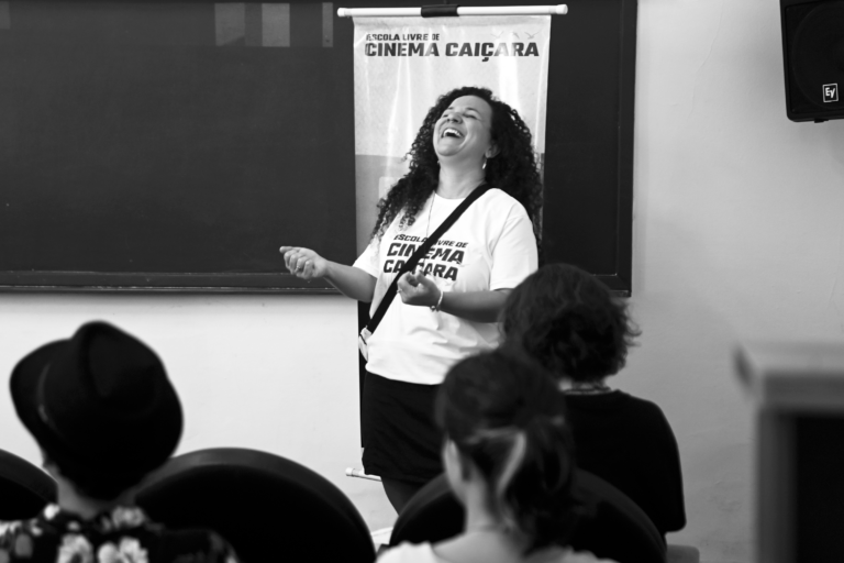 fotografia em preto e branco de uma mulher em frente a um grupo de pessoas que está sentado. Ela está de pé, gesticulando com uma das mãos, sorrindo e inclinando a cabeça para cima. A mulher está usando uma camiseta branca com um texto que "ESCOLA LIVRE DE CINEMA CAIÇARA". Ao fundo, há um quadro negro. Na parte inferior da imagem, há algumas pessoas sentadas, de costas para a câmera, assistindo à apresentação