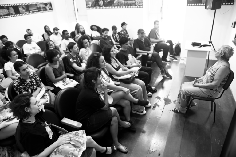 foto em preto e branco de uma sala de aula no formato auditório com várias pessoas jovens sentadas em cadeiras, são os alunos da Escola Livre de Cinema Caiçara assistindo a uma apresentação ou palestra. As pessoas no público estão sentadas em fileiras, algumas olhando para frente e outras para baixo, talvez lendo ou escrevendo. À frente, sentada, há uma mulher, de cabelos curtos e grisalhos, ela é palestrante. Ela fala olhando para o público.