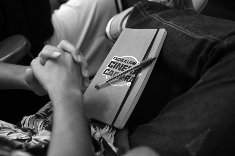 A imagem em preto e branco mostra uma pessoa sentada, com as mãos entrelaçadas sobre o colo. Ao lado das mãos, há um caderno com a capa escrita "ESCOLA DE CINEMA CAIÇARA" e uma caneta colocada sobre ele. A pessoa está vestindo uma roupa com estampa floral. Ao fundo, é possível ver parte de outra pessoa, mas está fora de foco.