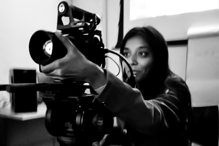 fotografia em preto e branco de uma mulher jovem segurando uma câmera de vídeo grande. A jovem é aluna da Escola de Cinema Caiçara. Ela sorri levemente e olha diretamente para a câmera e segura a lente da frente. O ambiente é interno