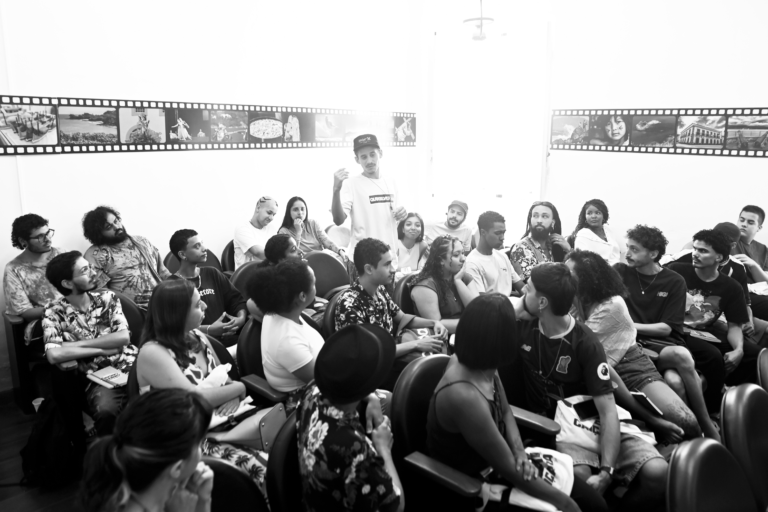 Na foto em preto e branco, um grupo de pessoas sentadas em cadeiras em uma sala de aula participando de uma discussão em grupo. A sala está cheia, com pessoas jovens e de estilos diferentes. São estudantes da Escola Livre de Cinema Caiçara dentro da sala de aula, olhando atentamente da direita para esquerda um aluno em pé falando em direção a eles.