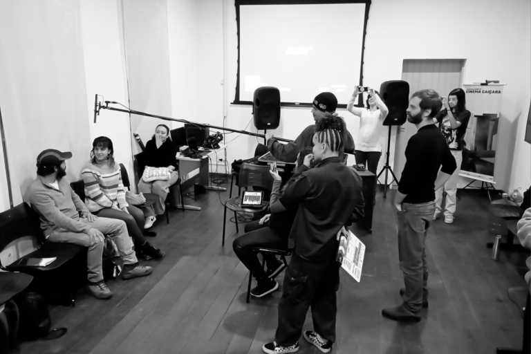 Foto em preto e branco de um grupo de pessoas em um ambiente interno, a sala da Escola de Cinema. Algumas pessoas estão sentadas em cadeiras, enquanto outras estão de pé no que parece ser um tipo de aula, com equipamentos de áudio visíveis, como microfones e alto-falantes. Um aluno em pé segurando um bastão longo com um microfone na ponta. Ele direciona o microfone para um aluno e uma aluna que estão sentados. Uma aluna ao lado observa a cena sentada com a mão no queixo. Atrás da cena, o professor observa em pé a atividade da turma. No canto direito da foto uma aluna com celular registra a cena e ao fundo uma tela de projeção.