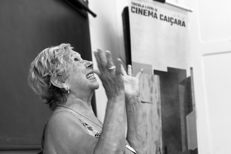 fotografia em preto e branco de uma mulher idosa, de perfil, gesticulando, apontando as palmas das mãos para cima, olhando para cima e sorrindo. Ela tem cabelos curtos e grisalhos. Ao lado, de fundo, há uma parede com um letreiro que diz "ESCOLA LIVRE DE CINEMA CAIÇARA".