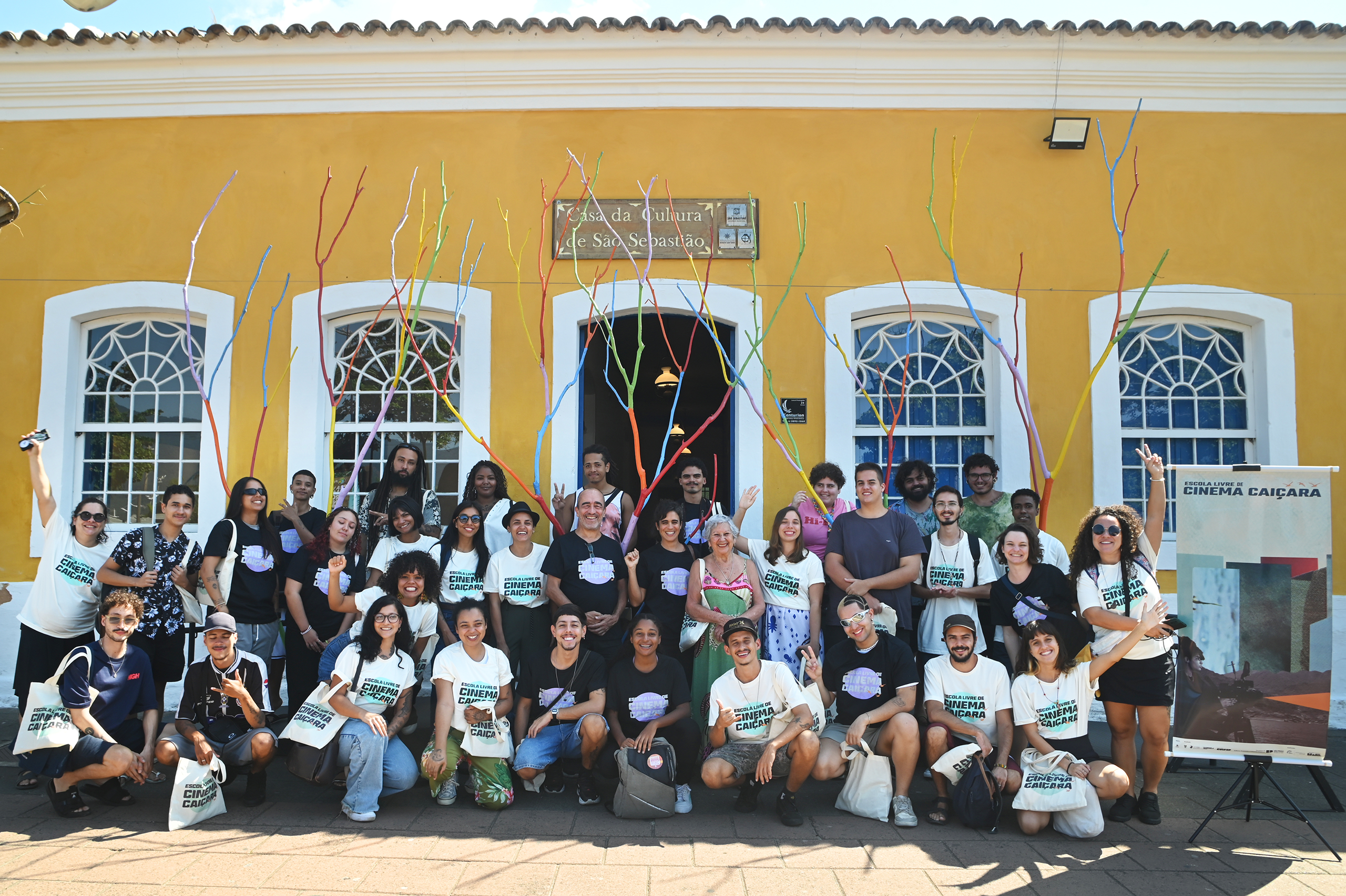 Foto com alunos e equipe da Escola Livre de Cinema Caiçara na aula de estreia da escola. Aproximadamente trinta e cinco pessoas, vestidos com camisetas e bolsas com o logotipo da escola, fazem gestos de positividade e sorriem, sentados e em pé de frente a um casarão antigo de cor amarela, quatro janelas grandes brancas e uma porta central aberta com um placa acima descrito "Casa de Cultura de São Sebastião. Ao lado direito um banner com o logotipo da escola pendurado em um tripé de metal.